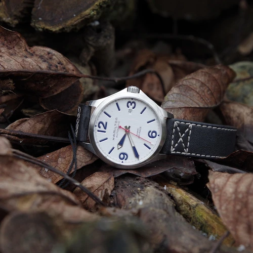 Reloj de campo (Field Watch) con esfera blanca y números arábigos grandes, sobre hojas secas, destacando su resistencia militar.